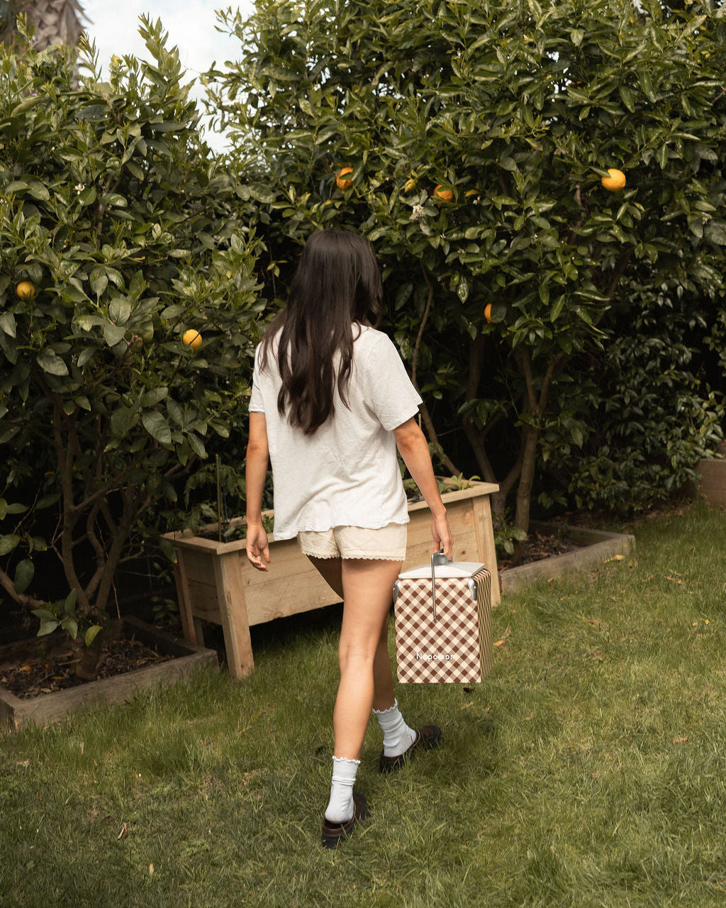 A woman seen from behind walking on a grassy lawn toward orange trees in a garden, holding a brown and white checkered cooler