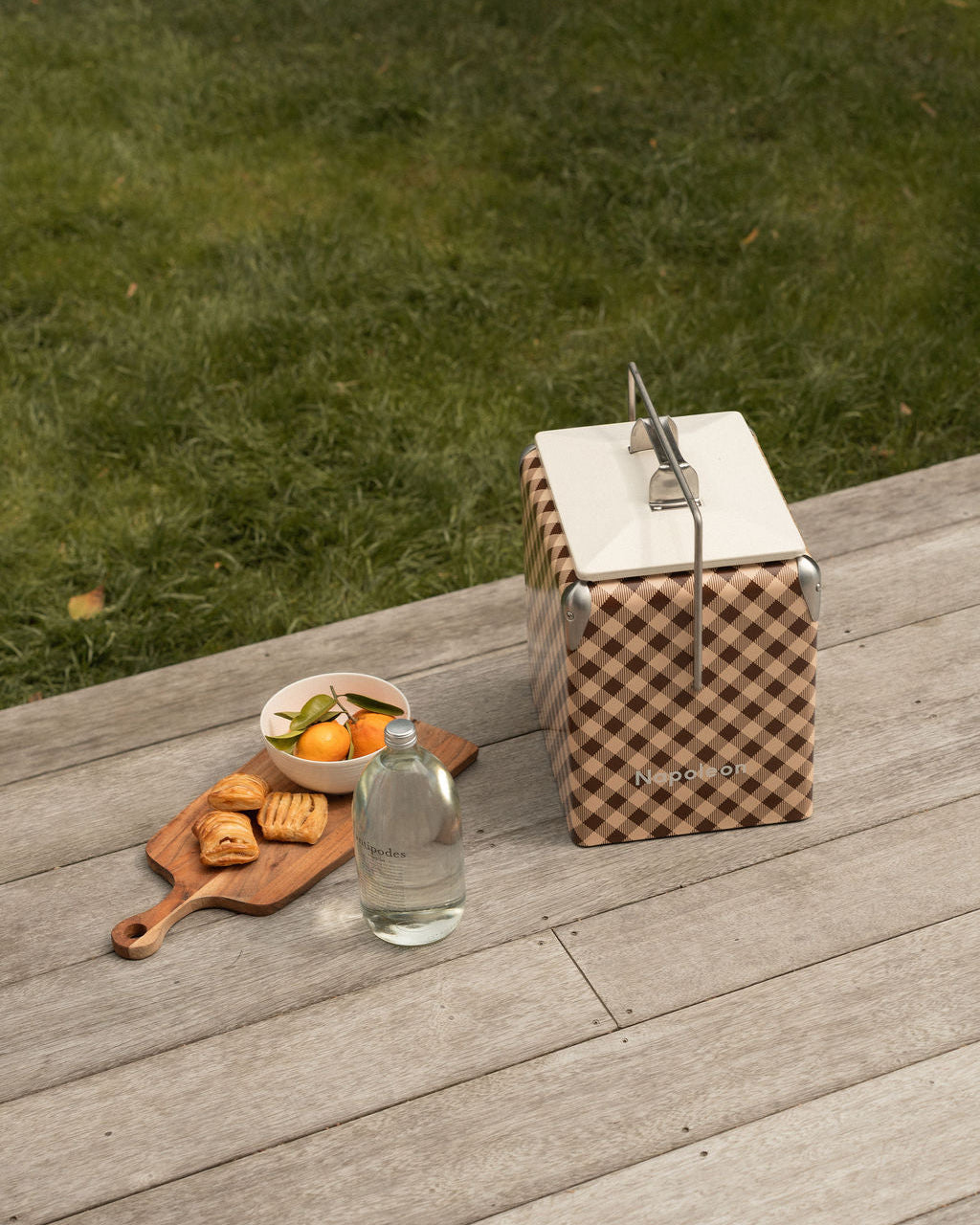 A picnic-style scene on a wooden deck with a brown-and-beige gingham cooler with sparkling water in a glass bottle fruit and pastry. green grass in background